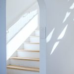 Bright minimalist interior featuring a white staircase with an arch and natural light casting shadows.