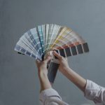 Hands displaying a fan of paint color swatches against a gray wall, ideal for home renovation projects.